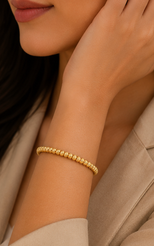 A close-up of a woman's wrist wearing a delicate gold beaded bracelet, with her hand resting near her face and a beige blazer visible in the background.