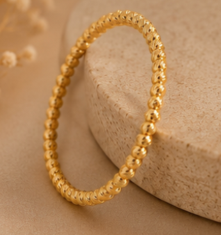 A gold beaded bracelet with a textured, pebble-like finish, displayed on a beige stone prop against a warm beige background.